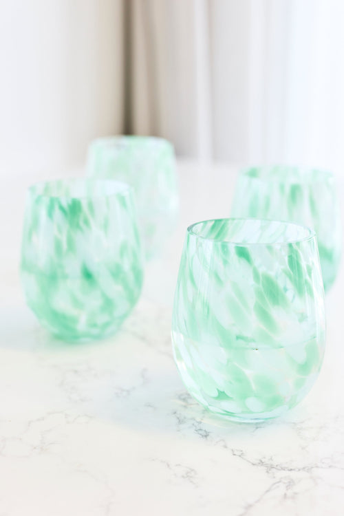 Four stemless wine glasses with a light green and white marbled pattern, arranged on a white marble surface with a soft-focus background.