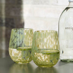 A clear glass bottle labeled 'antipodes' with two textured green drinking glasses on a round dark green side table, set against a light stone wall with a green chair partially visible on the left.