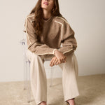 Woman sitting on a chair wearing a brown sweater and beige pants against a plain background