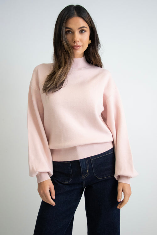 Woman wearing a soft blush knit jumper with balloon sleeves, a funnel neck, and dark denim jeans.