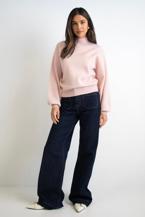 Woman wearing a soft blush knit jumper with balloon sleeves, a funnel neck, and dark denim jeans.