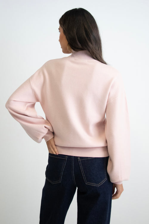 Woman wearing a soft blush knit jumper with balloon sleeves, a funnel neck, and dark denim jeans.