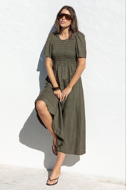A woman wearing an olive green midi dress and sunglasses stands next to a swimming pool in front of a white wall.