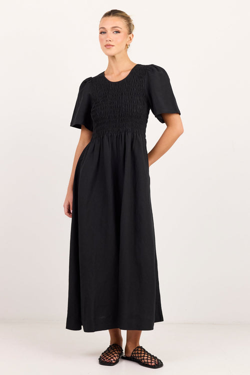 A woman modeling a black midi dress with short puff sleeves, a smocked bodice, and a gathered skirt, paired with black strappy flat sandals against a plain white studio background.