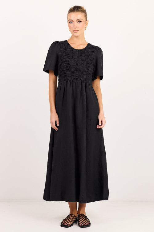 A woman modeling a black midi dress with short puff sleeves, a smocked bodice, and a gathered skirt, paired with black strappy flat sandals against a plain white studio background.
