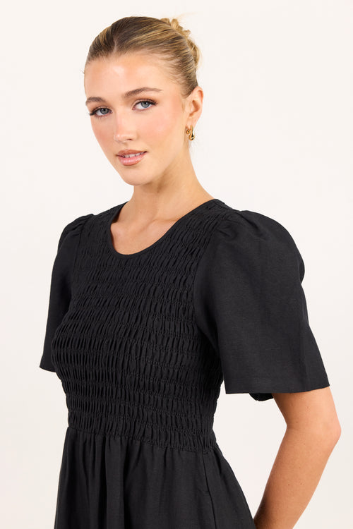 A woman modeling a black short-sleeve dress with a smocked bodice and puffed sleeves against a plain white studio background.