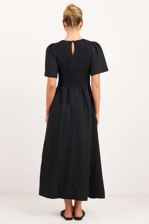 A woman viewed from the back wearing a black midi dress with short puff sleeves, a smocked bodice, and a small keyhole cutout at the back, standing against a plain white studio background.