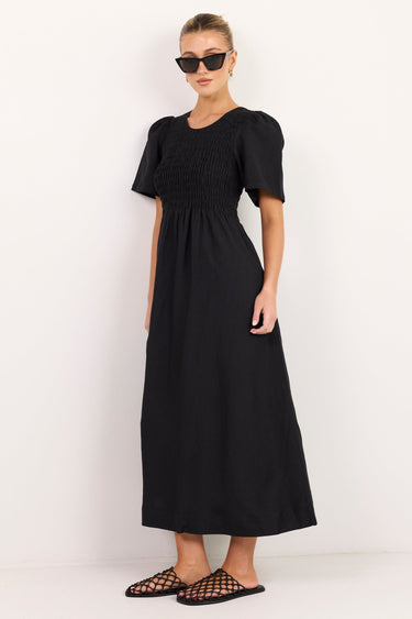 A woman modeling a black midi dress with short puff sleeves, a smocked bodice, and a gathered skirt, paired with black strappy flat sandals against a plain white studio background.