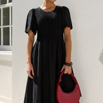 A woman wearing a black midi dress with puff sleeves and a smocked bodice, holding a red crescent-shaped handbag, standing against a white stucco wall.