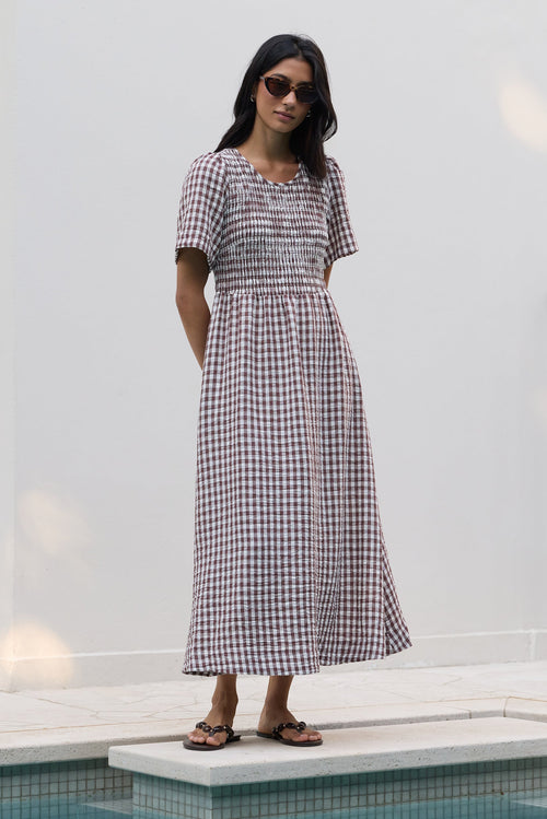 A woman wearing a short-sleeve gingham midi dress and sunglasses stands on a tiled pool deck against a white wall.