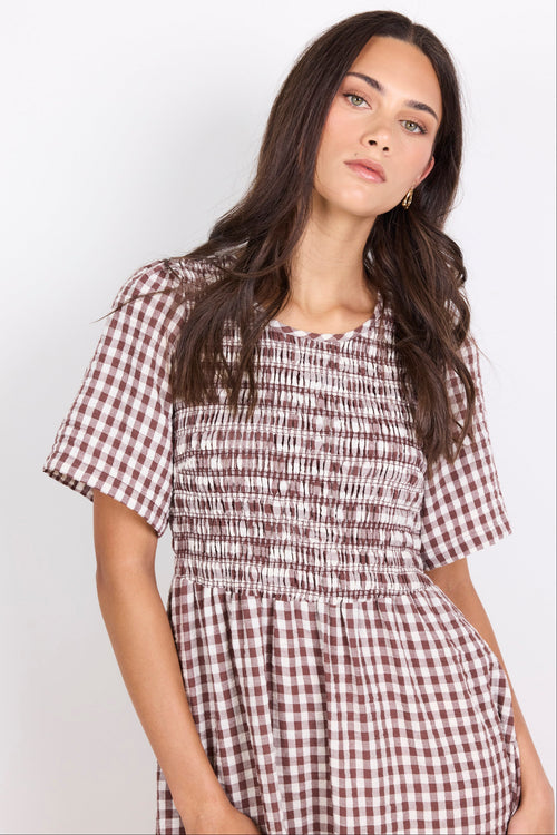 A woman wearing a short-sleeve smocked dress with a brown and white gingham pattern, photographed against a plain light gray studio background.
