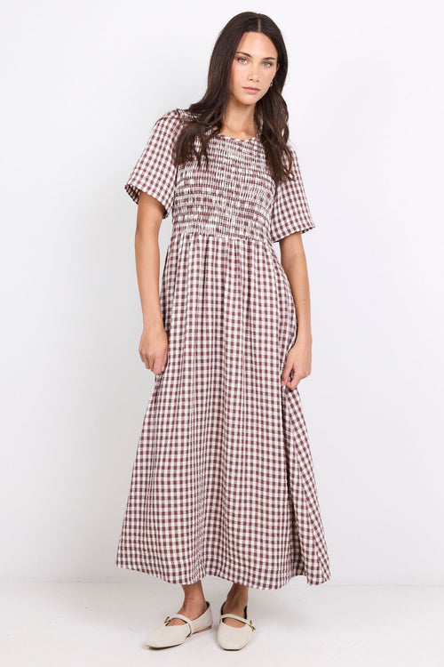 A woman modeling a short-sleeved, smocked-waist maxi dress in a brown and white gingham pattern, paired with white flat shoes, against a plain white studio background.