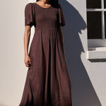 A woman wearing a dark brown midi dress with short flutter sleeves and a smocked bodice, standing outdoors against a white wall with a window.