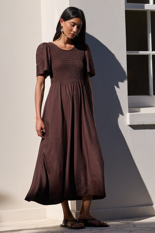 A woman wearing a dark brown midi dress with short flutter sleeves and a smocked bodice, standing outdoors against a white wall with a window.