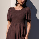 A woman wearing a dark brown smocked midi dress with short flutter sleeves, standing against a sunlit beige wall with a strong shadow.