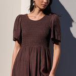 A woman wearing a dark brown smocked midi dress with short flutter sleeves, standing against a light-colored wall with strong sunlight and shadows.