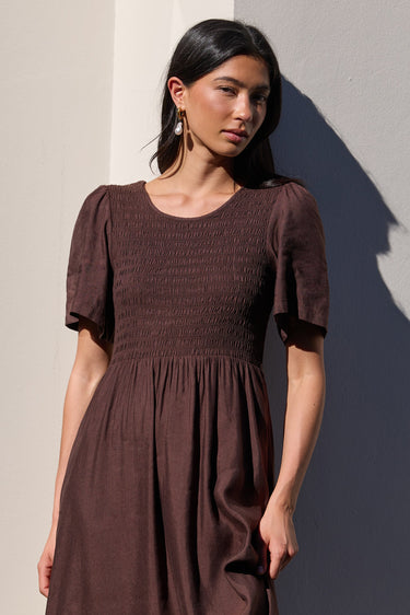 A woman wearing a dark brown smocked midi dress with short flutter sleeves, standing against a sunlit beige wall with a strong shadow.