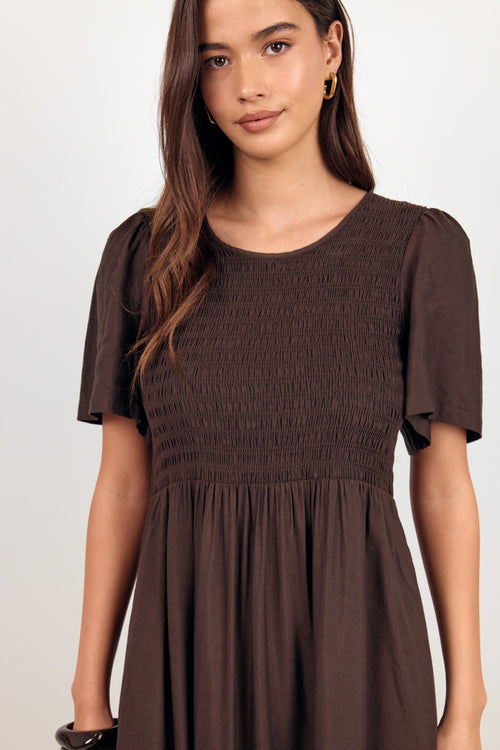 A woman wearing a dark brown smocked dress with short flutter sleeves and a gathered waist, photographed against a plain light background.