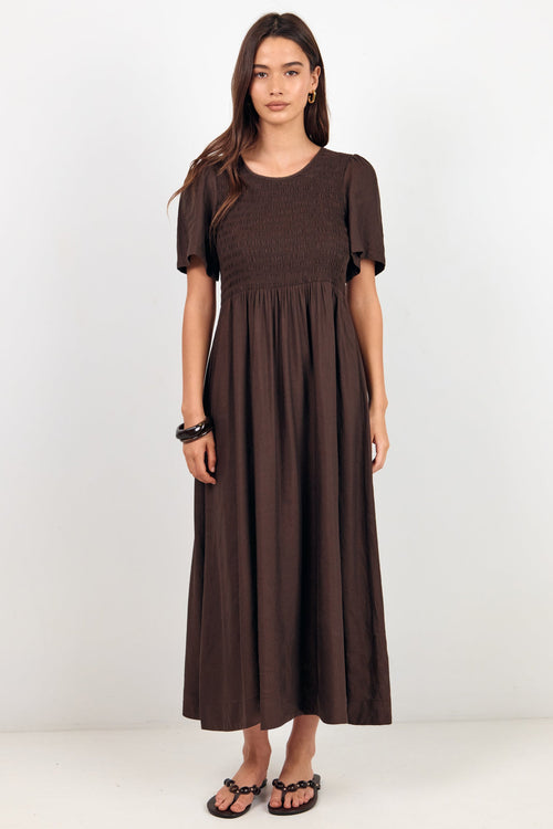 A woman modeling a dark brown short-sleeve maxi dress with a smocked bodice and gathered skirt, standing against a plain white studio background.