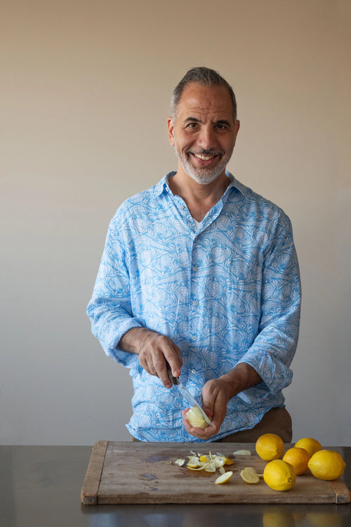 Man in a blue patterned shirt cutting lemons on a wooden board with a neutral background