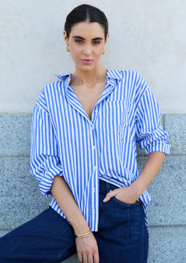 Woman wearing a blue and white striped shirt and dark jeans against a light-colored wall.