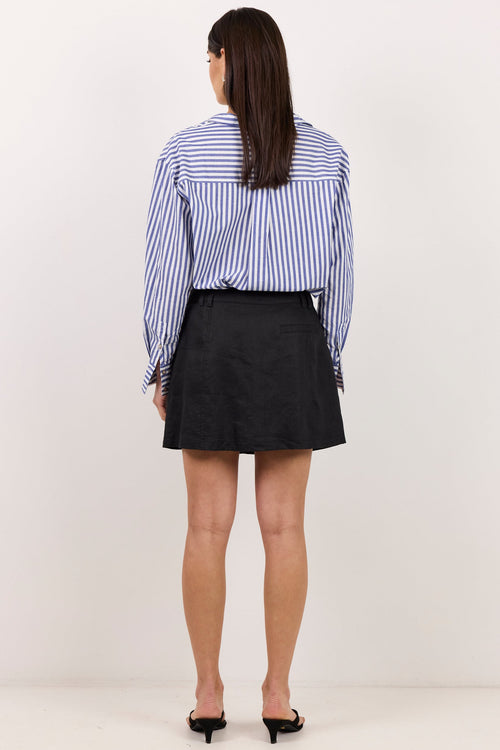 A woman viewed from behind wearing a blue and white vertically striped button-down shirt, a black mini skirt, and black pointed-toe flats, standing against a plain off-white studio background.