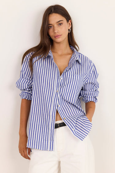 A woman modeling a blue and white vertically striped button-down shirt with white pants, standing against a plain off-white background.