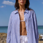 A woman stands on a sandy beach wearing a blue and white vertically striped button-up shirt, white drawstring pants, and dark sunglasses, with the ocean and blue sky in the background.