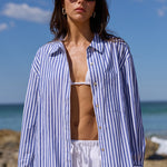 A woman stands on a sandy beach wearing an oversized blue and white vertically striped button-up shirt, white drawstring pants, and dark sunglasses, with the ocean and blue sky behind her.