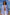 A woman stands on a sandy beach wearing an oversized blue and white vertically striped button-up shirt, white drawstring pants, and dark sunglasses, with the ocean and blue sky behind her.