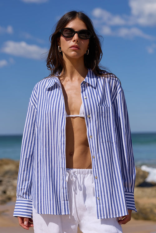 A woman stands on a sandy beach wearing an oversized blue and white vertically striped button-up shirt, white drawstring pants, and dark sunglasses, with the ocean and blue sky behind her.
