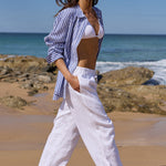 A woman walks barefoot on a sandy beach wearing a blue and white striped button-up shirt, white wide-leg pants, and a white bikini top under a blue sky with clouds.
