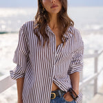 A woman wearing a brown and white vertically striped button-down shirt and blue denim jeans, standing on a sunny boardwalk with the ocean in the background.