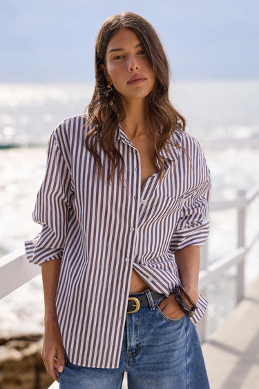 A woman wearing a brown and white vertically striped button-down shirt and blue denim jeans, standing on a sunny boardwalk with the ocean in the background.