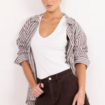 A woman modeling a casual outfit consisting of a white V-neck tank top, dark brown shorts, and a brown and white striped button-up shirt draped off her shoulders, set against a plain white background.