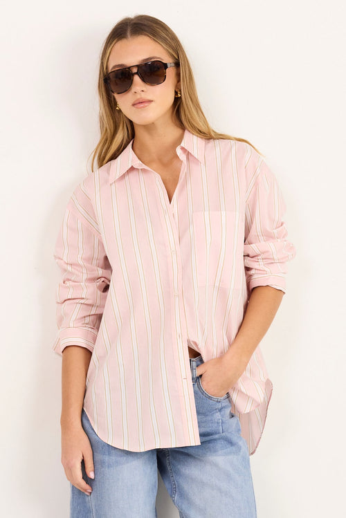 A woman modeling a light pink and white vertically striped button-down shirt, paired with light-wash blue jeans and white heeled sandals, standing against a plain white studio background.