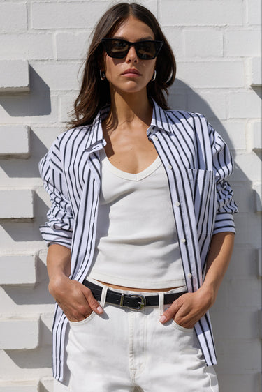 A woman wearing a navy and white vertically striped button-down shirt over a white tank top, paired with white pants and black sunglasses, posing against a white brick wall.