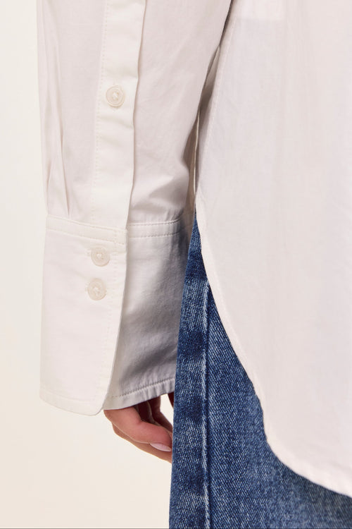 Close-up of a white long-sleeve button-up shirt worn with blue jeans, showing the sleeve cuff with two buttons and a vertical welt pocket.