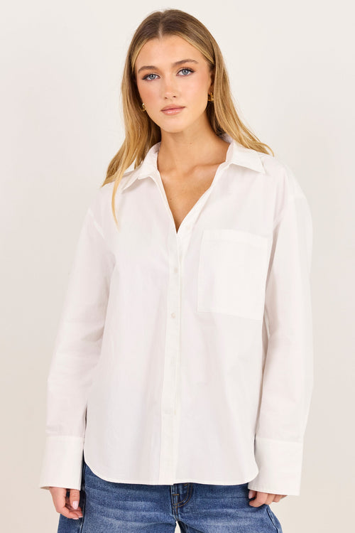 A woman modeling a white long-sleeve button-up shirt with a chest pocket, paired with blue jeans, against a plain off-white studio background.