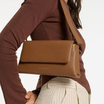 A structured camel-brown leather shoulder bag with a rectangular flap-over design and fine pebble texture. It features a detachable wide strap with black metal hardware and "Status Anxiety." subtly printed in white on the front base.