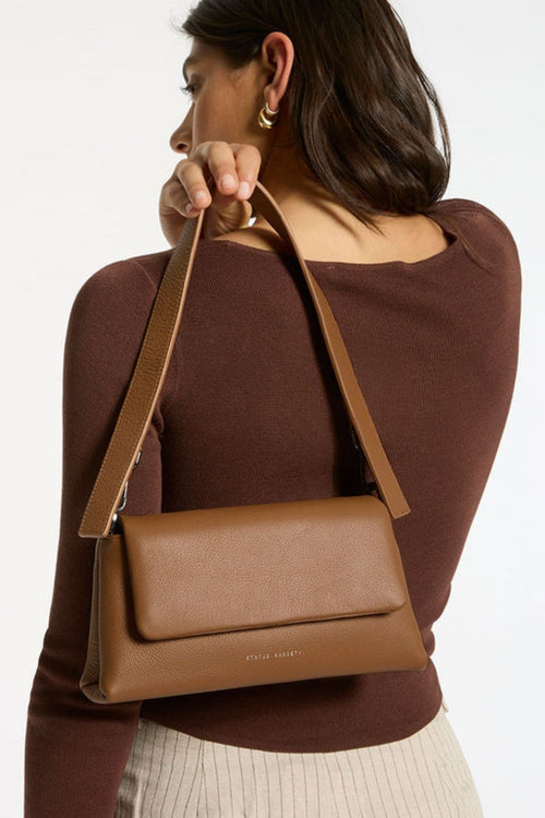 A structured camel-brown leather shoulder bag with a rectangular flap-over design and fine pebble texture. It features a detachable wide strap with black metal hardware and 