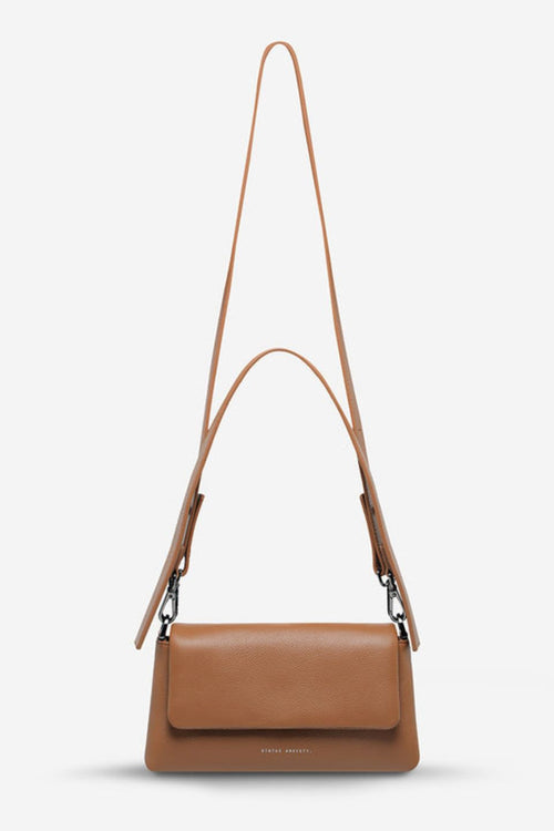 A structured camel-brown leather shoulder bag with a rectangular flap-over design and fine pebble texture. It features a detachable wide strap with black metal hardware and 