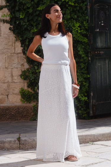 Woman in a white dress standing outdoors against a stone wall and greenery.