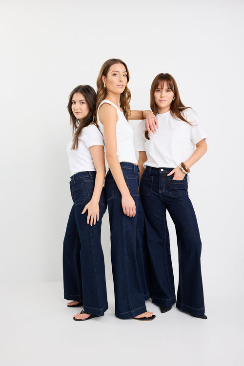 Three women wearing white tops and dark blue jeans standing together against a white background