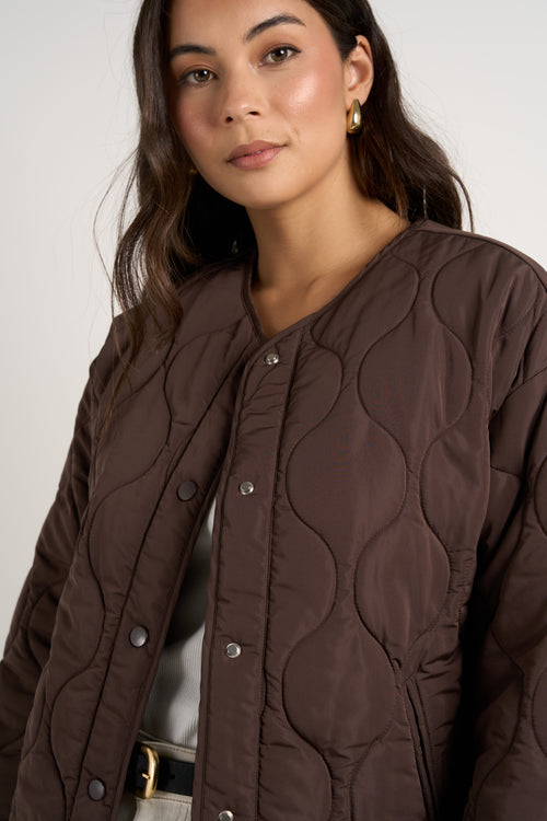 model wears a chocolate brown collarless puffer jacket with  cream jeans