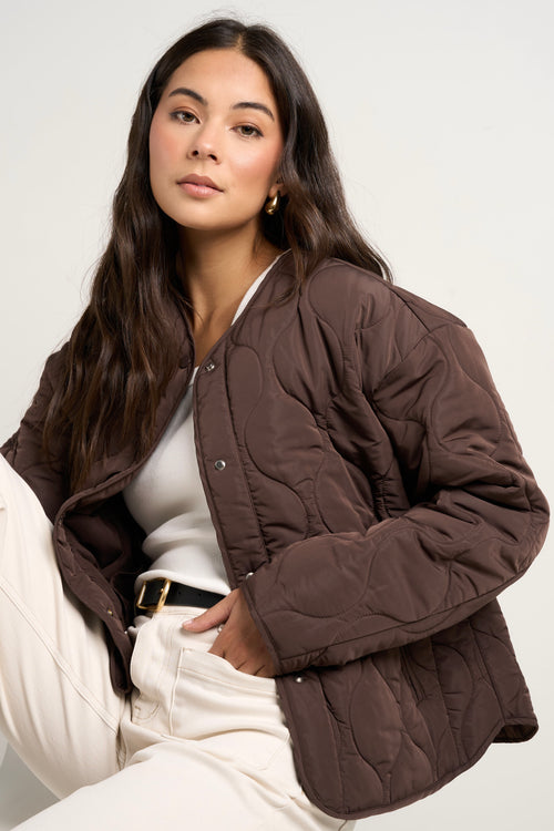 model wears a chocolate brown collarless puffer jacket with  cream jeans