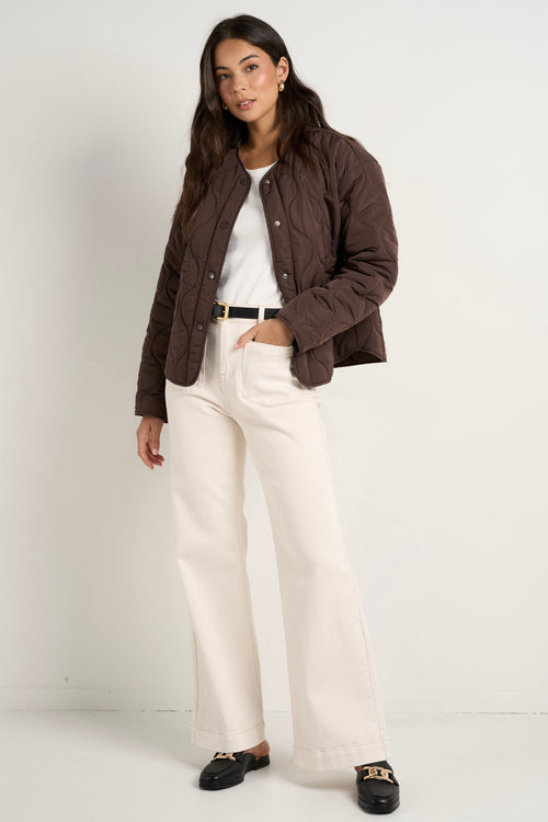 model wears a chocolate brown collarless puffer jacket with  cream jeans