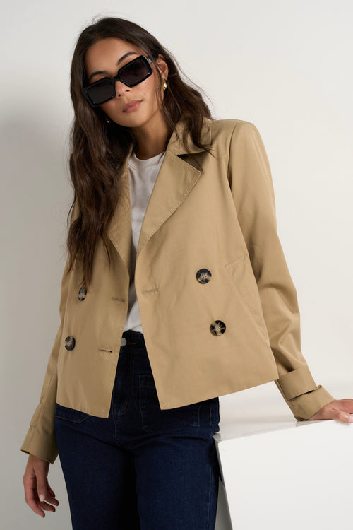 Model wears camel beige trench coat with dark indigo jeans