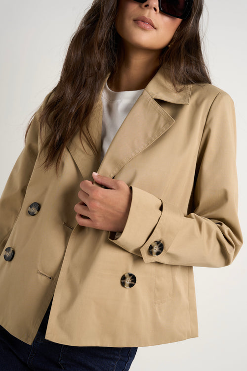 Model wears camel beige trench coat with dark indigo jeans