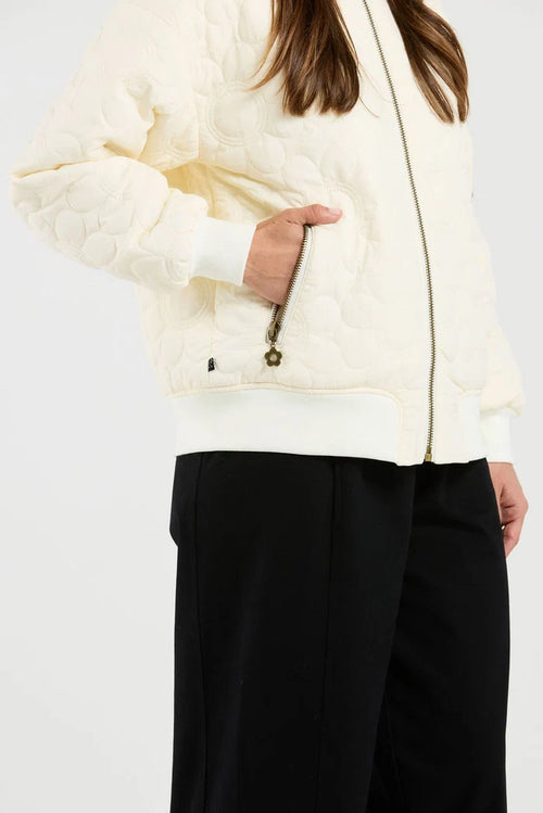A close-up of a person wearing a cream quilted zip-up jacket with ribbed cuffs and hem, paired with black pants, against a plain light background.